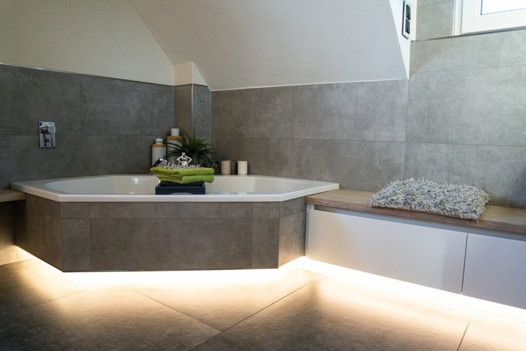 Moderne Eckbadewanne mit LED-Beleuchtung, grünen Handtüchern und gemütlicher Sitzbank – stilvolles Bad von KLU-BAD